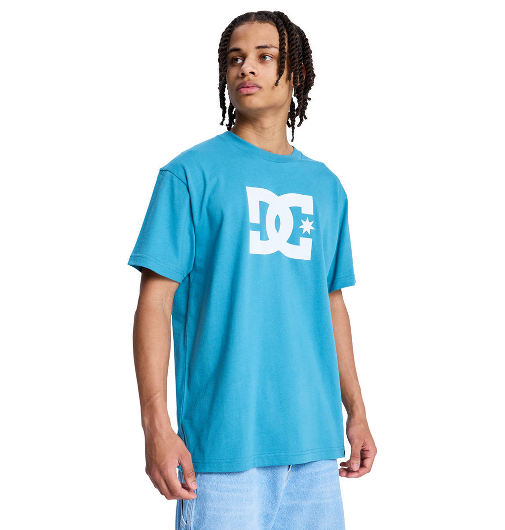 A person with braided hair wearing a bright blue Reebok DC Star Hss t-shirt and light blue jeans stands turned slightly to the side against a white background.