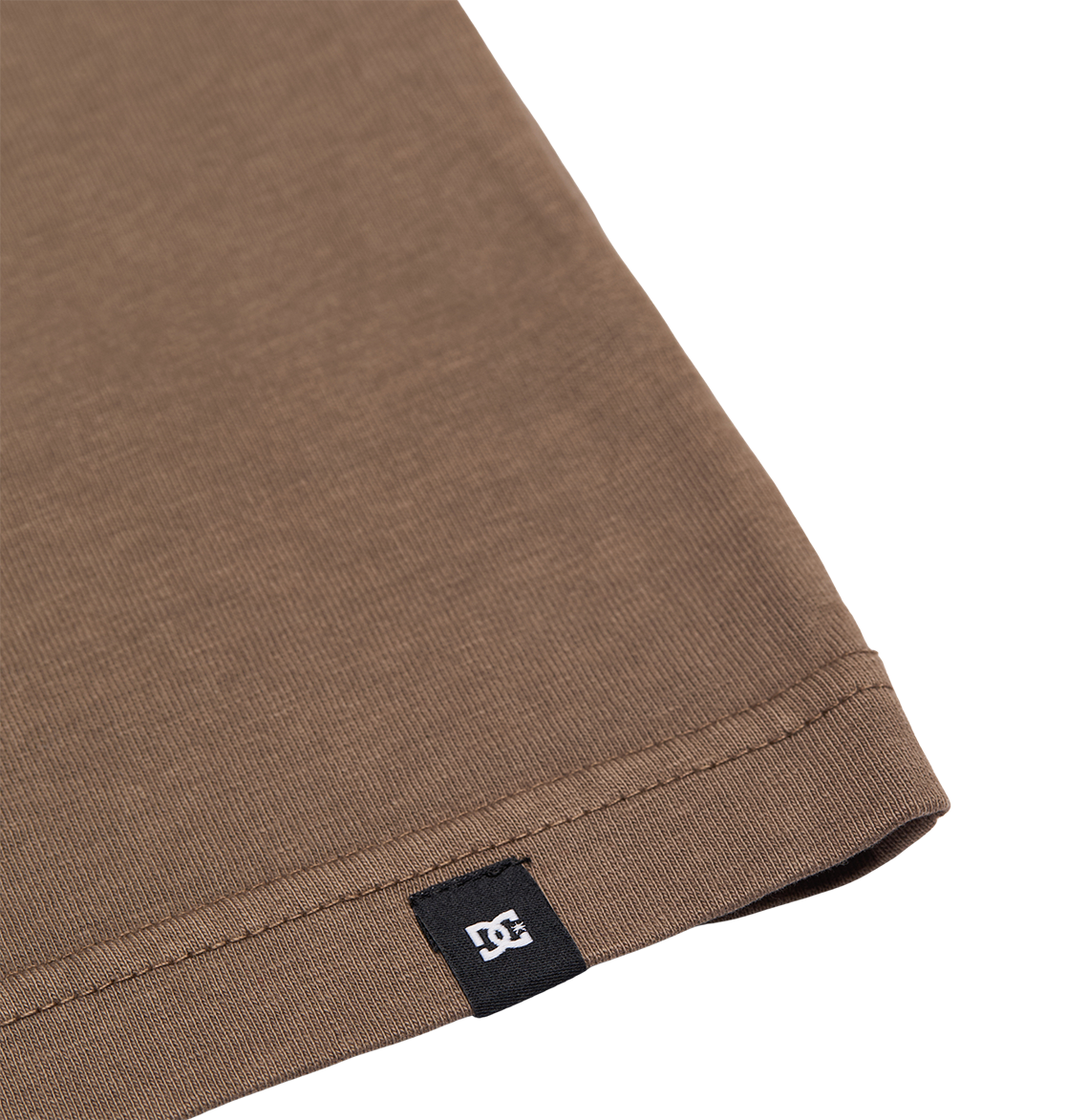 Close-up of the hem of the Men's DC Star Pigment Dye Tee by DC Shoes in brown, featuring a small black tag with a white DC logo sewn into the seam. The background is plain white.