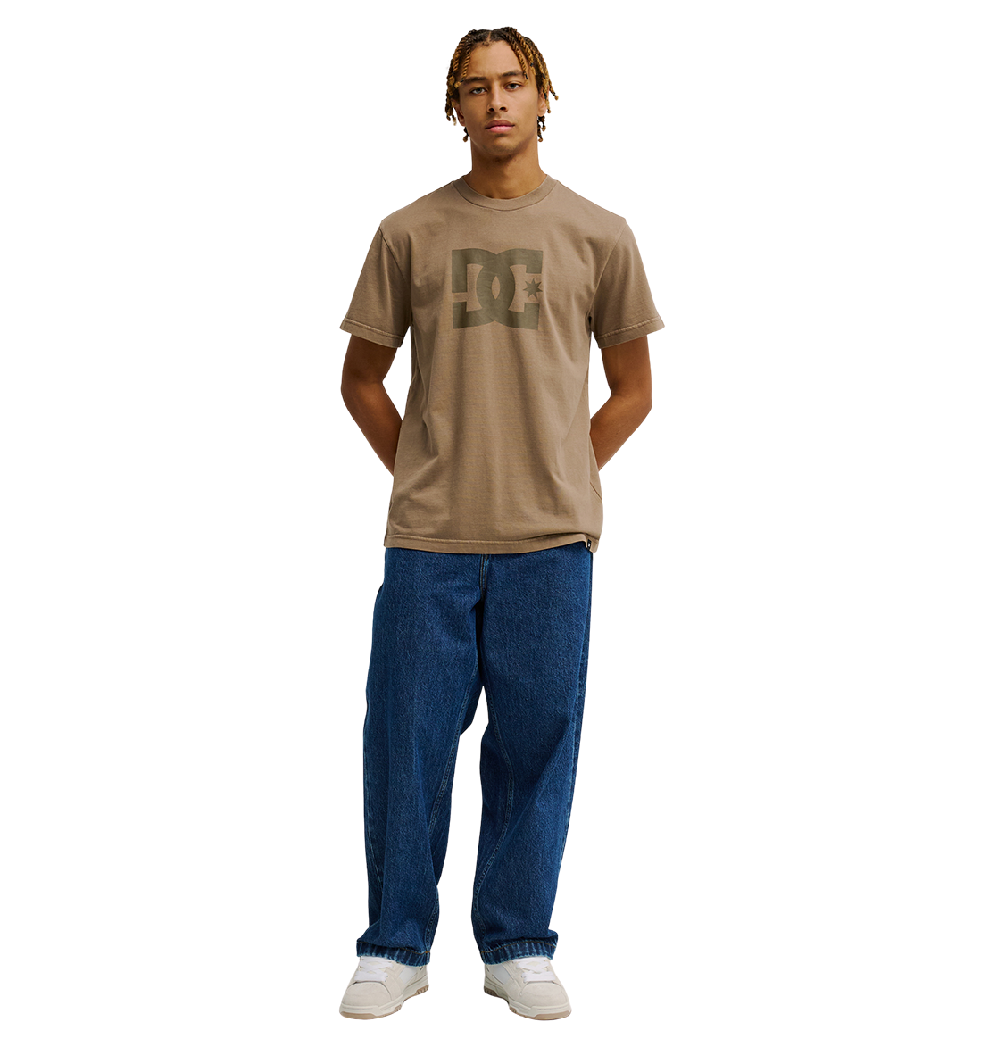 A person with short, blonde braids wears the Men's DC Star Pigment Dye Tee from DC Shoes—a relaxed fit brown skate t-shirt featuring iconic DC branding—paired with loose blue jeans and white sneakers, standing with hands behind their back against a plain white background.