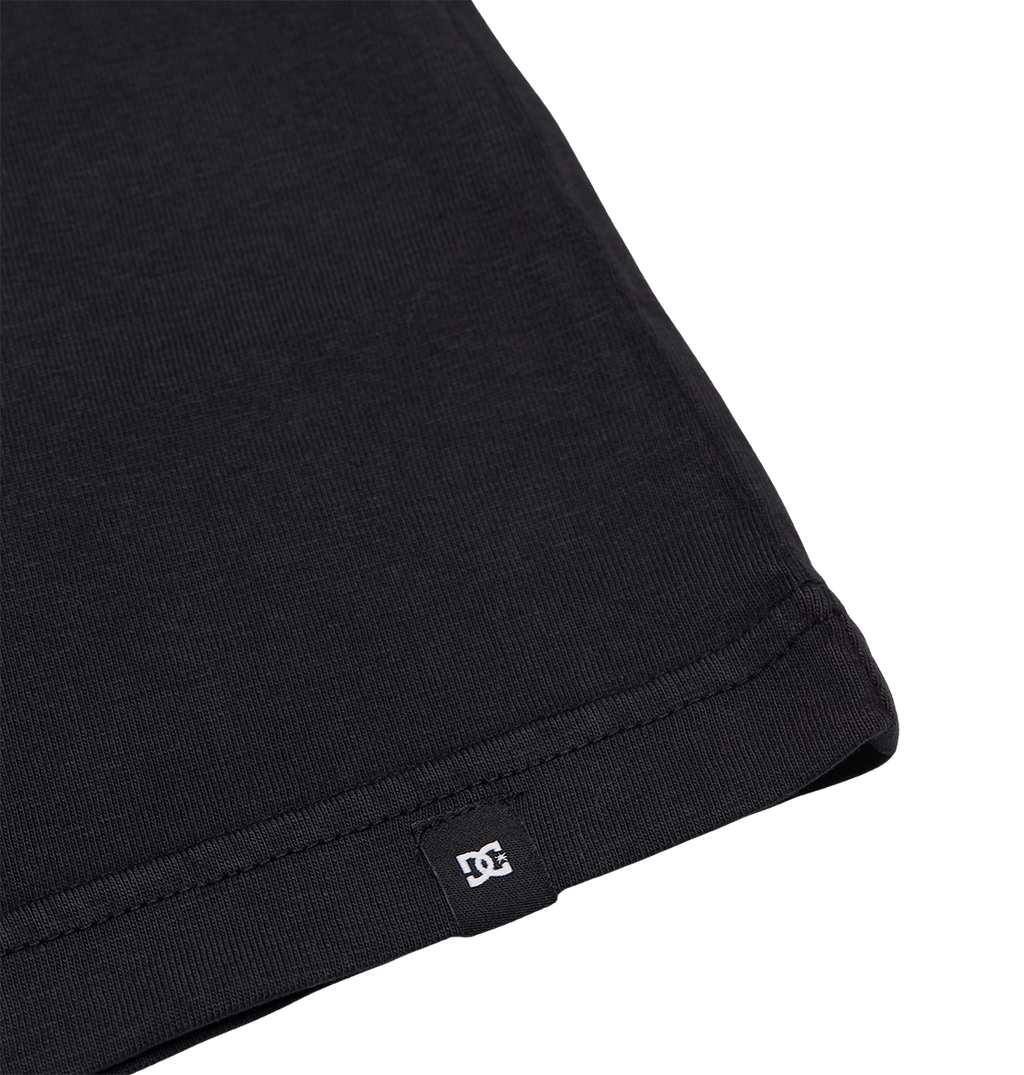 Close-up of the bottom hem of the DC Shoes Speratic Tee Youth skateboarding shirt in black, featuring a small black tag with a white DC Shoes logo, set against a plain white background.