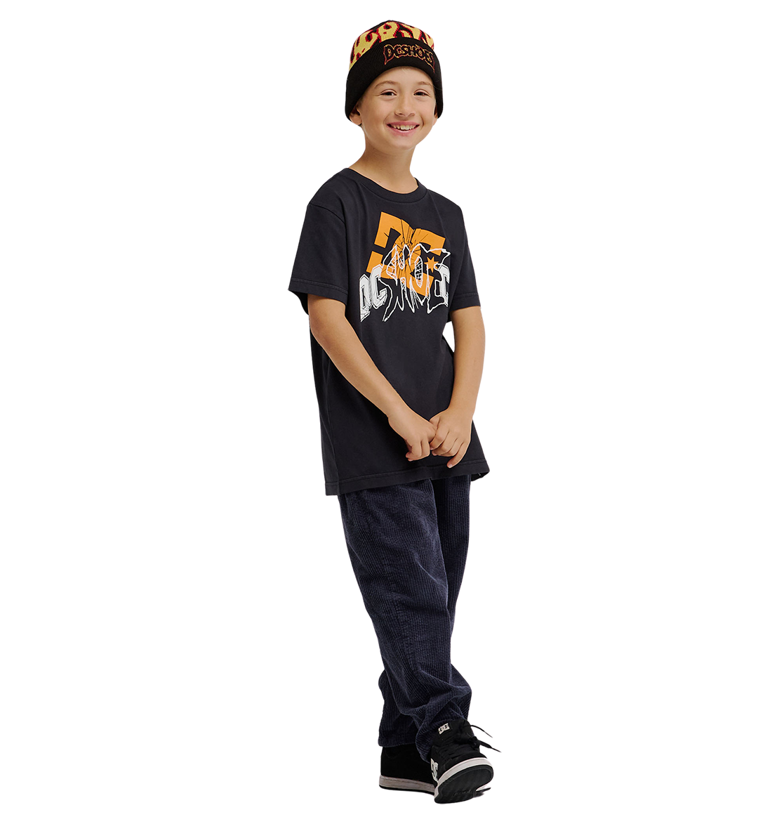 A smiling child stands against a plain white background wearing the DC Shoes DC Speratic Tee Youth in black, paired with dark pants, black sneakers, and a black beanie with orange lettering.