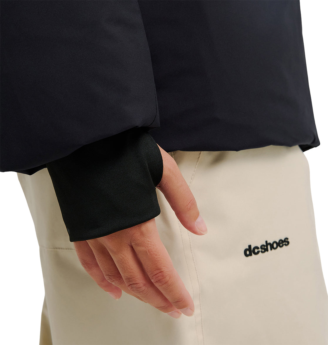 A close-up of a person’s hand, partially covered by the sleeve—with thumb hole—of the DC Shoes Aura Puffer Jacket in black, resting on cream-colored pants featuring the DC Shoes logo.