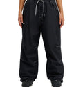 A person is shown from the waist down, standing against a white background, wearing loose-fitting DC Shoes Trackstar Pants 10K in black with white stitching, a drawstring waist, cargo pockets, and paired with black shoes.